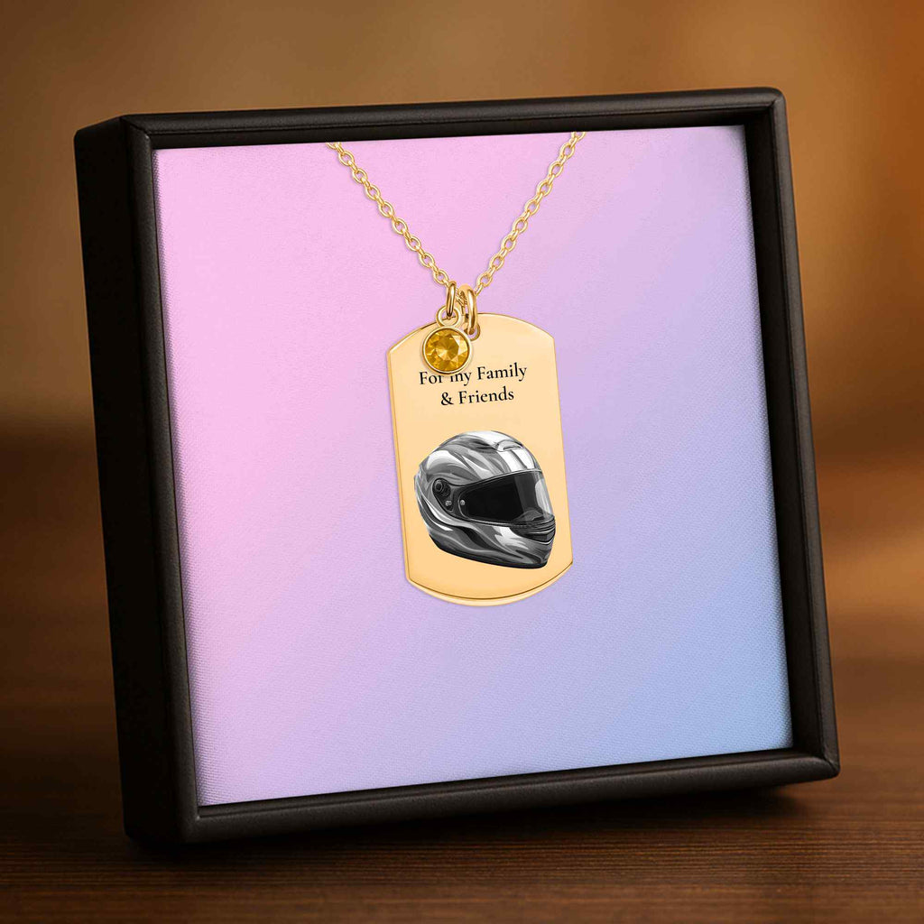 A gold-tone dog tag necklace with a small charm and printed text, displayed inside a black presentation box with a pink-to-blue gradient insert. The dog tag features a motorcycle helmet graphic and the text 'For my Family & Friends'.