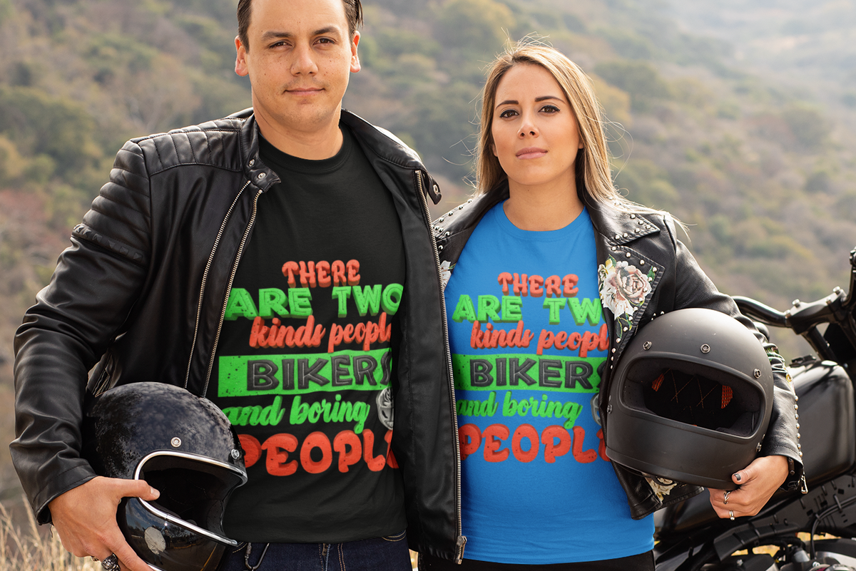 A man and a woman stand outdoors in front of a motorcycle on a hillside, wearing matching graphic T-shirts with the text 'THERE ARE TWO kinda people BIKERS and boring PEOPLE'. The man wears a black T-shirt under a black leather jacket and holds a helmet, while the woman wears a bright blue T-shirt under a black leather jacket and holds a black helmet.