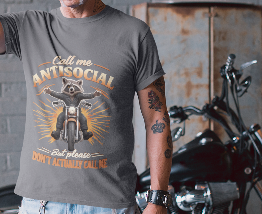 A man wearing a dark gray graphic t-shirt with a motorcycle design, light wash ripped jeans, and a black bandana stands in front of a black motorcycle in a garage setting.