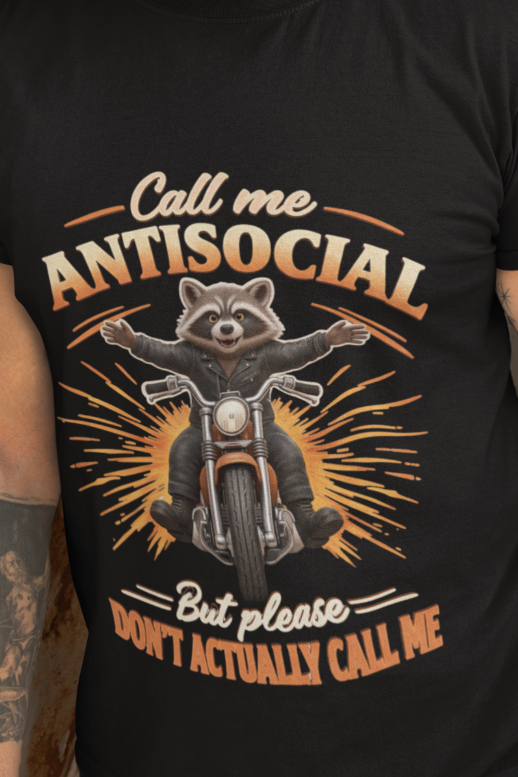 A man wearing a black graphic t-shirt with a raccoon on a motorcycle and the text 'Call me ANTISocial But please DON'T actually call me', standing in front of a rusted metal door.