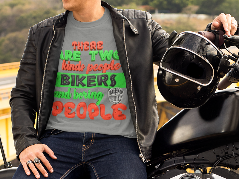 A man sits on a black cruiser motorcycle outdoors beside a bridge, wearing a graphic T-shirt, black leather jacket, jeans, and brown boots, with a black helmet resting on the motorcycle.