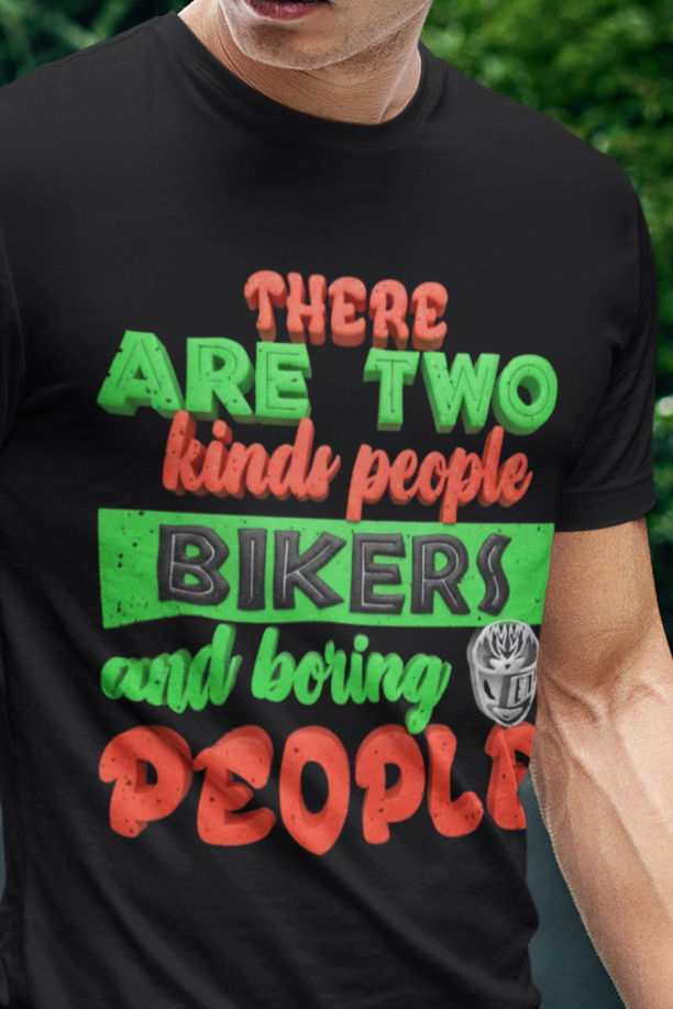 A man wearing a black graphic t-shirt with the text 'THERE ARE TWO kinds people BIKERS and boring PEOPLE' in green and orange, paired with blue jeans, standing outdoors on a grassy lawn with a modern building and greenery in the background.