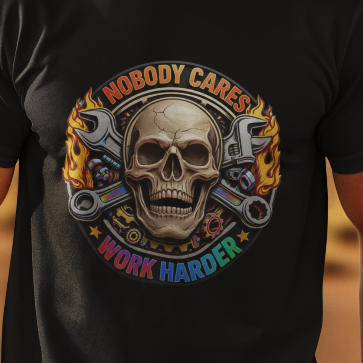 A man wearing a black graphic t-shirt with a skull and wrenches design that reads 'NOBODY CARES WORK HARDER', standing in a desert landscape with rock formations in the background.